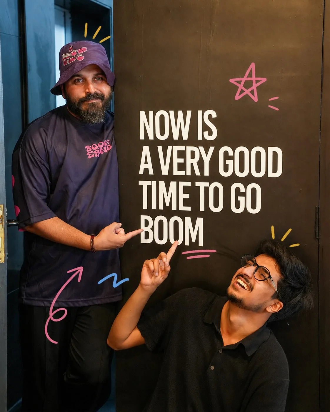 Two Boomerang staff at the 'Now is a very good time to go boom' wall, one in the tee and hat